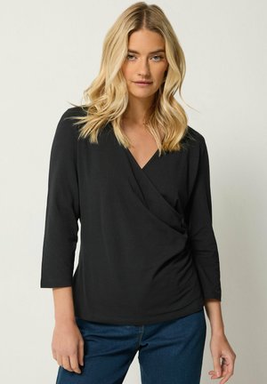 Blonde Frau trägt ein schwarzes, langärmliges Wickeltop und blaue Jeans und steht vor einem einfarbigen, hellen Hintergrund.