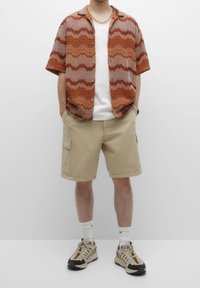 Chemise à manches courtes à boutons avec un motif en zigzag orange et marron, associée à un short cargo beige et des chaussures de sport beiges.