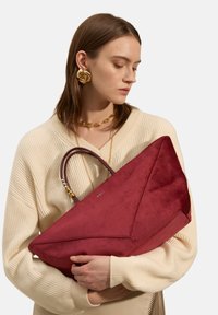 Sac fourre-tout en daim rouge avec un design angulaire, présentant une surface texturée et deux accents de poignée, tenu contre un pull en tricot clair.