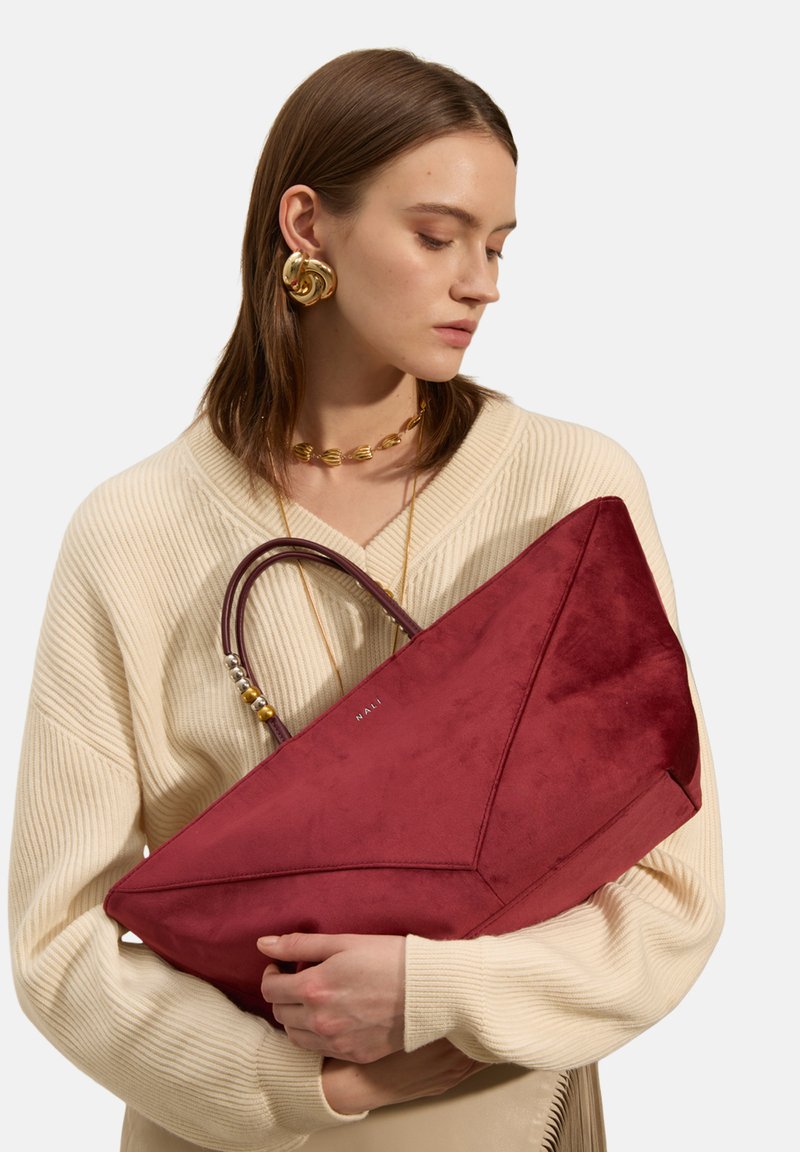 Sac fourre-tout en daim rouge avec un design angulaire, présentant une surface texturée et deux accents de poignée, tenu contre un pull en tricot clair.