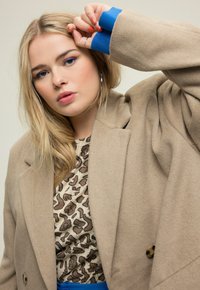 Blazer de lana beige con grandes solapas, blusa de estampado de leopardo marrón debajo y detalles en azul en los puños; textura suave, detalles de botones visibles.