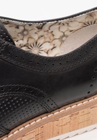 Chaussure en cuir noir avec des détails perforés, une doublure intérieure à motif floral et une semelle texturée en liège avec une couture blanche.