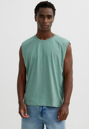 Hombre con cabello corto y rizado, vestido con una camiseta sin mangas verde y vaqueros azules, de pie frente a un fondo liso.