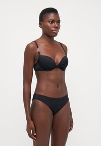 Černý bikini s designem zkrouceného předního dílu, měkké košíčky, nastavitelné ramínka a stříbrné kovové detaily. Hladká, elastická tkanina s minimálním pokrytím.
