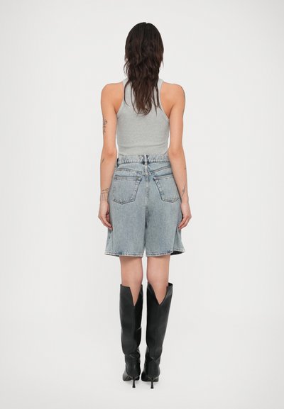 Γυναίκα με μακριά σκουρόχρωμα μαλλιά, ντυμένη με γκρι αμάνικη μπλούζα, ανοιχτόχρωμα τζιν σορτς και μαύρες ψηλοτάκουνες μπότες μέχρι το γόνατο, που στέκεται μπροστά σε λευκό φόντο.