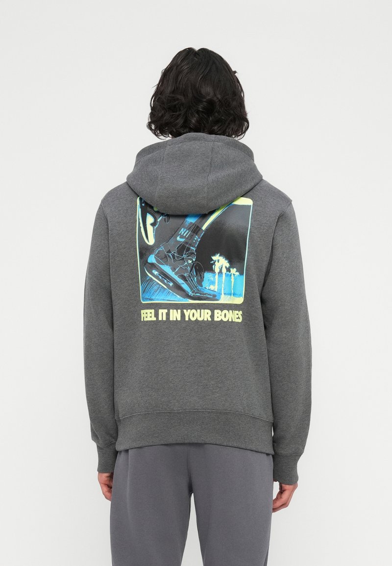 Pessoa com cabelo escuro usa um hoodie cinza com uma estampa neon de um ténis e palmeiras, com o texto "SENTE NO TEU CORPO" nas costas.