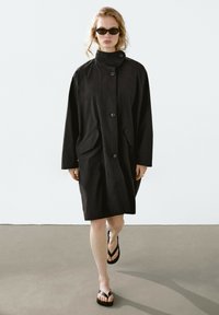 Černý dlouhý trench coat s vysokým límcem, předními knoflíky a dvěma velkými kapsami. Nosený přes žabky, s volným, rovný designem.