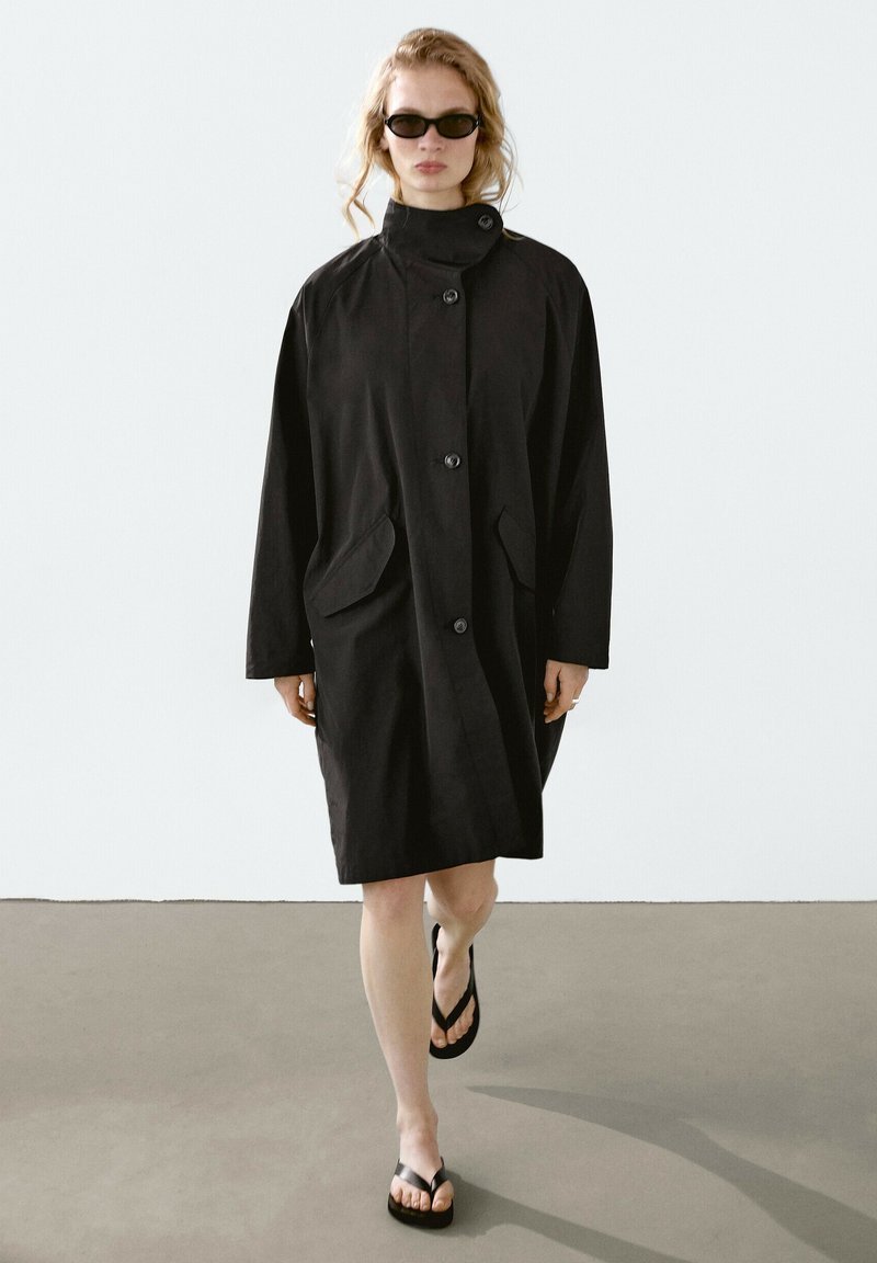 Černý dlouhý trench coat s vysokým límcem, předními knoflíky a dvěma velkými kapsami. Nosený přes žabky, s volným, rovný designem.