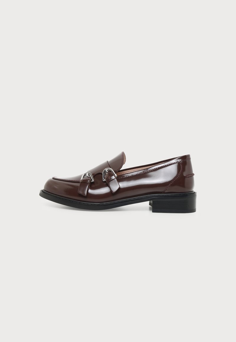 Mocassin en cuir marron foncé brillant avec double boucles argentées sur semelle noire à talon bas sur fond clair uni.