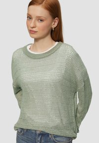 Hellgrüner, gestrickter Pullover mit Rundhalsausschnitt, strukturiertem Muster und lockerer Passform. Über einem weißen Oberteil getragen, das aus einem weichen, leichten Stoff besteht.