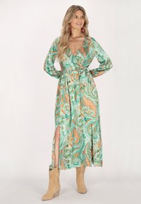 Rochie înfășurată verde cu imprimeu paisley, cu mâneci lungi, decolteu adânc în V, talie strânsă și fante pe lateral, accentuată cu motive portocalii și teal. Pur tată cu cizme bej.
