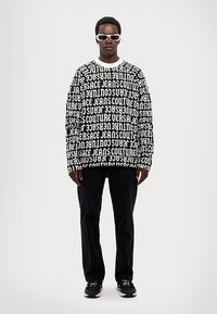 Přes oversized černobílá grafická mikina s opakujícím se textem. V kombinaci s tmavými kalhotami a černými teniskami. Model má na sobě bílé sluneční brýle.