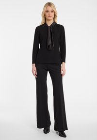 Schwarzes Langarmoberteil mit einem Satinband am Kragen, kombiniert mit weiten schwarzen Hosen, die subtile Funken enthalten. Beide Kleidungsstücke haben eine glatte Textur.