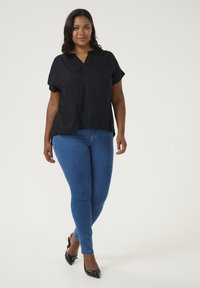 Blouse noire à manches courtes avec décolleté en V, associée à un jean bleu ajusté et des escarpins à talons aiguilles noirs sur un fond clair.