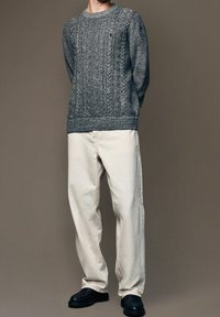 Suéter de punto gris con un patrón texturizado, combinado con pantalones de corte relajado en beige claro y zapatos gruesos negros, todo ello frente a un fondo neutro.