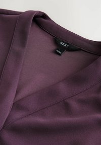 Fialový top s výstřihem do V z hladké látky, má jednoduchý design a minimální detail švů. Štítek nese nápis "NEXT" na černé značce.