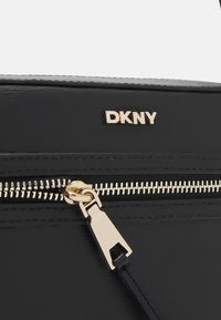 Black leather handbag with a shiny finish, featuring a gold metal logo, gold zipper, and a sleek design. Accents include a decorative zipper pull.