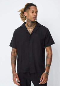 Chemise noire à manches courtes avec un col, en tissu texturé et dotée d'une petite poche poitrine.