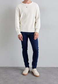 Biały prążkowany sweter dziany, ciemnoniebieskie dżinsy oraz jasne trampki z gumowymi podeszwami. Prosty design z luźnym fasonem i okrągłym dekoltem.