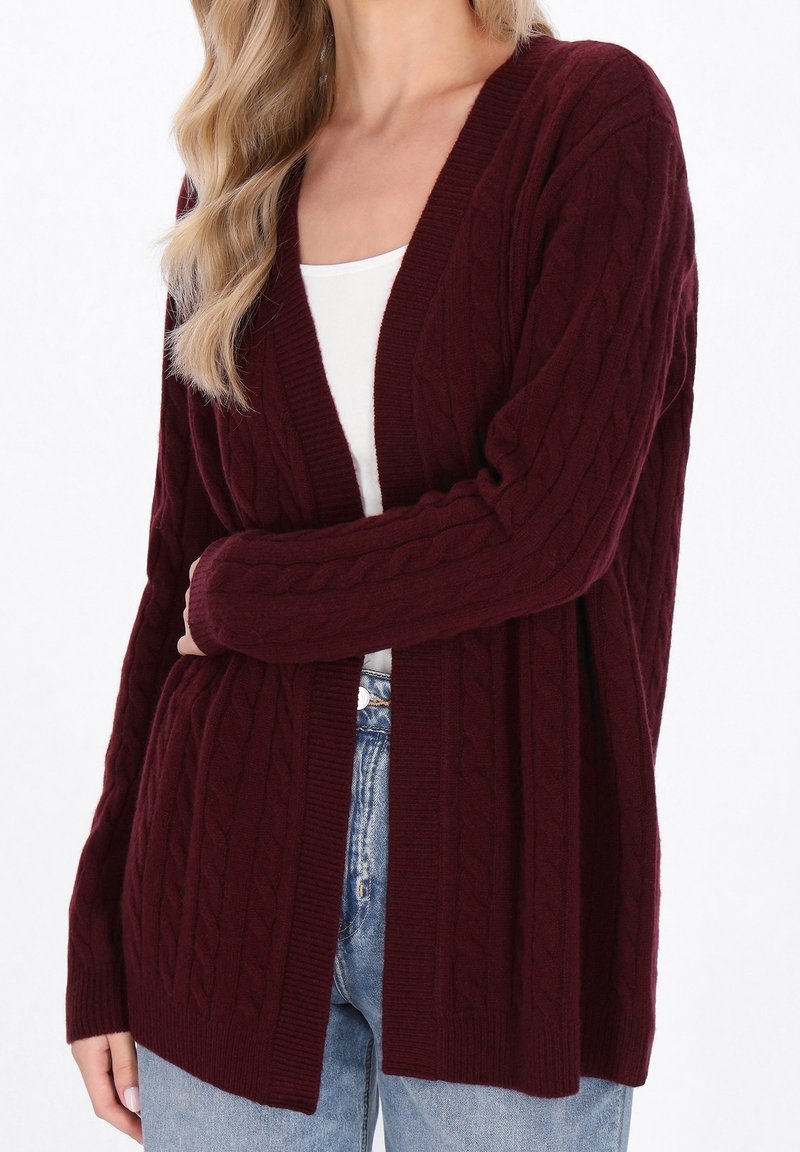 Femme portant un cardigan bordeaux à torsades sur un haut blanc et un jean bleu clair, debout avec un bras croisé.