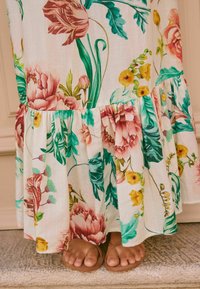 Robe maxi florale avec une jupe évasée, ornée de motifs roses, jaunes et verts sur fond blanc, agrémentée d'un ourlet volanté.