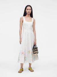 Femme portant une robe blanche sans manches avec une broderie florale colorée, tenant un sac à main rayé tissé, et portant des sandales vertes à bout ouvert.
