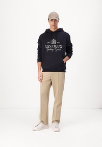 Felpa blu marina con tasca frontale e testo bianco; pantaloni beige a gamba dritta; sneakers bianche; cappellino color beige. Il modello è in piedi davanti a uno sfondo neutro.