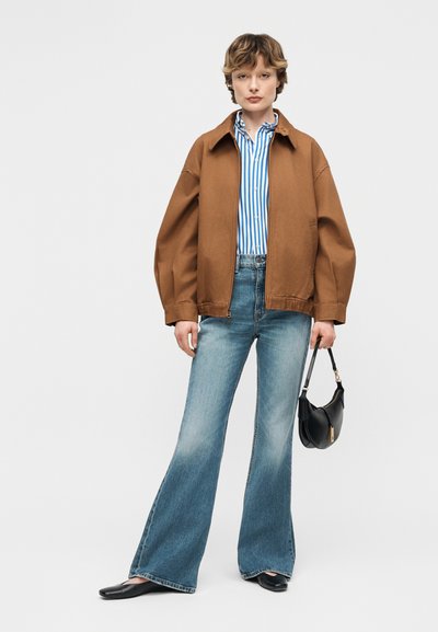 Νεαρό άτομο που φορά καφέ μπουφάν, ριγέ μπλε και λευκό πουκάμισο, καμπάνα μπλε τζιν, μαύρα φλατ παπούτσια, κρατώντας μικρή μαύρη τσάντα.