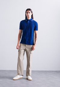 Tânăr cu păr lung și închis la culoare, îmbrăcat cu un tricou polo bleu închis, pantaloni cargo bej și adidași albi, stând pe un podea gri, împotriva unui perete alb.