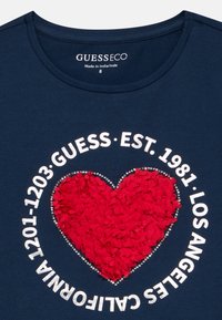 Μπλε ναυτικό πουκάμισο με μια υφή κόκκινης καρδιάς στο κέντρο, περιβαλλόμενο από λευκό κείμενο: «GUESS EST. 1981 LOS ANGELES CALIFORNIA 1201-1203.»