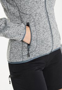 Grå texturerad fleecejacka med kontrasterande blå blixtlås, sidofickor och åtsittande ärmavslut, kombinerad med svarta byxor med blixtlås.