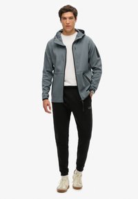 Chaqueta gris con capucha, cierre de cremallera, bolsillos frontales y detalles en negro, combinada con pantalones de chándal negros y zapatillas de color claro.