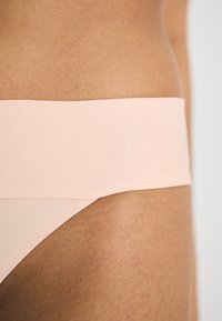 String rose clair avec une large ceinture lisse arborant un logo embossé discret. Le tissu semble doux et extensible.