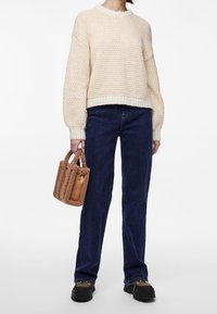 Krämfärgad tröja med en strukturerad stickad design, kombinerad med mörkblå rakbenade jeans. Håller en brun mini-väska i sydd päls. Tjocka svarta skor.