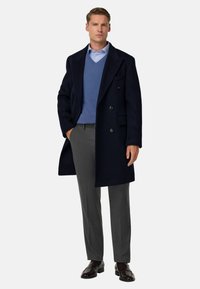 Navy modrý vlněný kabát se zdvojeným zapínáním, nošený přes světle modrý svetr a šedé kalhoty, doplněný hnědými koženými botami.