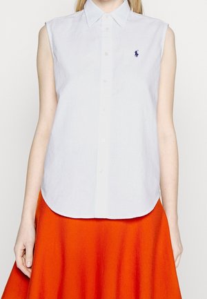 Mujer que lleva una camisa blanca sin mangas con cuello y un pequeño logo azul marino, combinada con una falda naranja brillante.