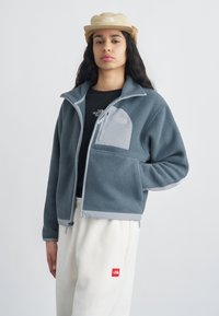 Νέος άνθρωπος που φοράει μπεζ καπέλο, γκρι φλις μπουφάν, μαύρο πουκάμισο και λευκό παντελόνι με λογότυπα της The North Face, στέκεται με το ένα χέρι στην τσέπη.