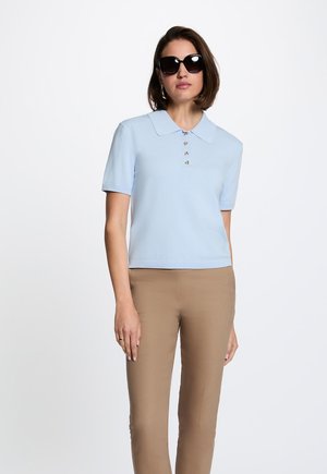 Kvinde iført sorte solbriller, lyseblå kortærmet poloshirt med knapper og beige bukser, stående foran en hvid baggrund.
