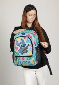 Sac à dos turquoise avec des imprimés floraux colorés et le personnage Stitch. Comprend une poche zippée à l'avant, des poches latérales en maille et des bretelles réglables.