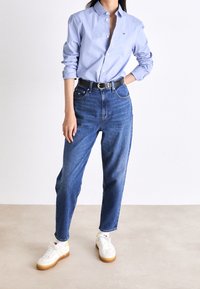 Camisa de botón azul claro de manga larga, combinada con jeans de cintura alta azules y zapatillas blancas con detalles amarillos. Cinturón negro visible.