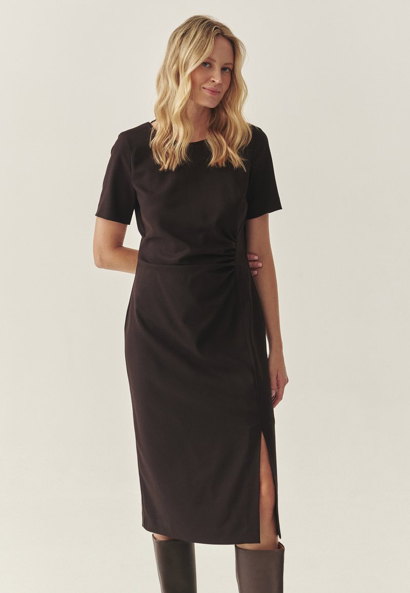 Vestido midi negro con mangas cortas, textura suave, abertura lateral y diseño ajustado. Combinado con botas negras hasta la rodilla. Silueta simple y elegante.