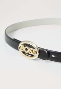 Black leather belt with a gold oval buckle featuring the embossed word "BOSS." Smooth texture, slim design, and holes for size adjustment.