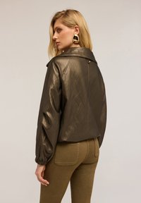 Giubbotto in pelle verde oliva metallico con colletto strutturato, polsini elasticizzati e chiusura lampo sulla schiena, abbinato a pantaloni aderenti verde oliva.