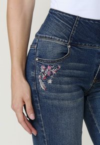 Jeans de mezclilla azul oscuro de talle alto, con bordados florales en rosa y blanco en el bolsillo trasero, de textura suave y corte clásico.