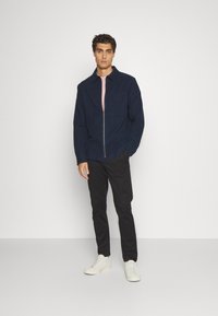 Giacca zip-up blu navy con colletto e taschino sul petto, indossata sopra una camicia rosa chiaro, abbinata a pantaloni neri e sneaker bianche.