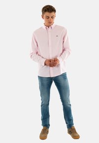 Camisa de botones de color rosa claro con cuello, combinada con jeans azules y zapatillas marrones. La camisa presenta un pequeño logo en el pecho.