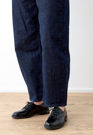 Personne portant un jean bleu foncé et des chaussures en cuir noir ciré à lacets, debout sur un sol en bois clair.