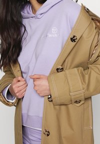 Gabardina beige con grandes botones, superpuesta a una sudadera con capucha de color púrpura claro. La gabardina presenta un pespunte blanco contrastante.