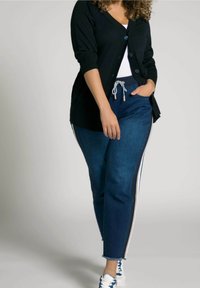 Cardigan negro con cierre de botones, camiseta blanca, joggers de mezclilla azules con rayas laterales y cordón, terminando con zapatillas blancas.