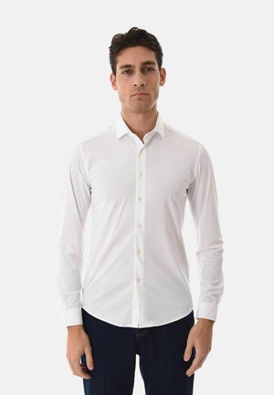 Uomo che indossa una camicia bianca aderente a maniche lunghe con bottoni e pantaloni scuri, in piedi contro uno sfondo chiaro e uniforme.